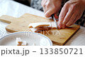 Person in their kitchen carefully slices pork belly on a wooden cutting board. Prepared pork pieces are on a plate nearby. Looks like they are prepping for a tasty meal. 133850721