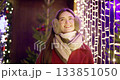 A young beautiful girl in a red coat walks in the evening at the European Christmas Market against the backdrop of illuminations and garlands. 133851050