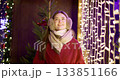 A young beautiful girl in a red coat walks in the evening at the European Christmas Market against the backdrop of illuminations and garlands. 133851166