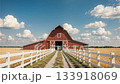 Idyllic red barn, dirt road, white fences, and golden wheat fields under a blue sky with clouds. A peaceful rural scene ideal for agriculture and country living. 133918069