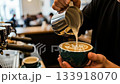 Close-up of barista hands pouring steamed milk into a ceramic cup, creating intricate latte art in a modern coffee shop. 133918070