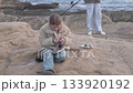 A young girl sits on a rocky coastline enjoying a small meal while her family fishes nearby. Dressed 133920192