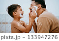 Father and son sharing a playful moment in the bathroom as the child applies shaving cream to his father's face, both laughing and enjoying the bonding experience 133947276