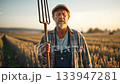 Senior farmer standing confidently in golden wheat field during sunset, gripping pitchfork and representing agricultural wisdom, resilience of farming lifestyle 133947281