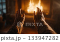 Woman wearing knitted sleeves warming her hands near crackling fireplace, experiencing serene winter evening with soft glowing flames casting warm shadows across rustic interior 133947282