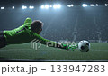 Athletic goalkeeper wearing a green uniform dives across the field, attempting to catch a soccer ball in a brightly lit stadium at night during a rainy match 133947283