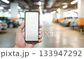 Professional holding smartphone with pristine white screen, displaying mockup in softly blurred parking garage environment, representing modern digital communication and technological connectivity 133947292