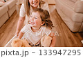 Loving mother playfully pushing laughing daughter resting with cuddly teddy bears inside wooden floored living room, sharing spontaneous moment of familial joy and connection 133947295