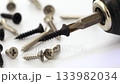 Close-up of a screwdriver with a black screw head, rotating at low speed, surrounded by various screws on a white surface. 133982034