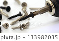 Close-up of a screwdriver with a black screw head, rotating at high speed, surrounded by various screws on a white surface. 133982035