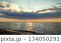 Seascape. Dark rainy clouds in blue orange sky over sea. Sunbeams from behind clouds. Sea waves on sand of seashore on sunset sunrise. Yellow reflection of sunlight on surface of sea waves. 134050291