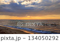 Sea waves with blue water on sand of seashore on sunset and sunrise. Yellow reflection of sunlight on surface of sea waves. Orange sky with dark rain clouds, horizon line and sea waves on sand. 134050292