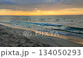 Sea waves with blue water on sand of seashore on sunset and sunrise. Yellow reflection of sunlight on surface of sea waves. Orange sky with dark rain clouds, horizon line and sea waves on sand. 134050293