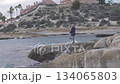 Quiet Shoreline Scene With Fishing And Hills, Calm Ocean View With Solitary Fisherman On Rocky Coast 134065803