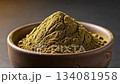 A perfectly sculpted pile of deep olive green matcha tea powder is presented in a brown ceramic bowl, highlighting its fine texture and vibrant color in a studio setting. 134081958