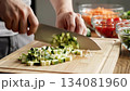 A chef hands precisely dice fresh green zucchini into small cubes on a wooden cutting board, with other colorful vegetables blurred in the background, highlighting healthy food preparation. 134081960