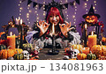 A young woman dressed in a playful gothic witch costume with red hair makes various hand gestures around a lit candle, surrounded by festive halloween decorations and pumpkins. 134081963