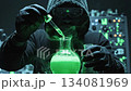 A mysterious hooded figure in gloves adds drops from a pipette into a round bottom flask containing a glowing green chemical solution that is bubbling and releasing smoke in a dark laboratory. 134081969