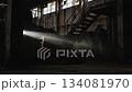 A dramatic beam of light cuts through the dusty air inside an abandoned industrial building, revealing old stairs, wooden crates, and a metal barrel in a dark, atmospheric interior. 134081970