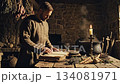 A somber man dressed in a historical tunic studies an ancient book on a rustic wooden table illuminated by candlelight in a dimly lit stone room with herbs and a steaming cauldron. 134081971