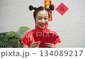 Happy young Asian woman in traditional red Cheongsam dress holding Ang Pao red envelopes for Chinese New Year celebration against white brick background Happy young Asian woman in traditional red Cheongsam dress holding Ang Pao red envelopes for Chinese New Year celebration against white brick background 134089217