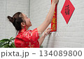 Happy young Asian woman wearing traditional red cheongsam dress decorating home with lucky couplets banner for Chinese New Year festival celebration Happy young Asian woman wearing traditional red cheongsam dress decorating home with lucky couplets banner for Chinese New Year festival celebration 134089800