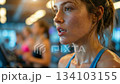 Exhausted woman with sweat on face breathes heavily during intense workout while woman standing in gym recovers; tired woman looks forward after hard fitness cardio training routine 134103155