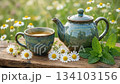 Steaming herbal tea in ceramic cup sits on wooden board while tea pot stands near fresh chamomile; hot tea with mint leaves creates relaxing morning atmosphere in sunny summer flower meadow. 134103156