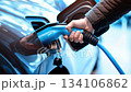 Close-up of man’s hand plugging in electric vehicle charger. Shiny blue car surface reflects ambient light. Concept of clean energy, EV technology, green transportation. Modern detail. Banner. 134106862