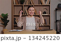 Positive Caucasian woman celebrating success holding smartphone raising hands with joy. Female smiling widely feeling excited. Sitting at wooden desk in home office with laptop and organized bookshelf 134245220