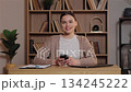 Caucasian woman holding smartphone with both hands and smiling at camera. Female sitting at wooden desk in home office taking break from work surrounded by bookshelves and laptop nearby. 134245222