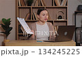 Mad Caucasian woman angrily pointing at paper with charts during video call on laptop. Female shouting at coworkers while discussing work mistakes. Sitting at wooden desk in home with bookshelves. 134245225