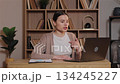 Caucasian woman gesturing and talking during video call on laptop. Female arguing with someone online expressing emotions while sitting at wooden desk in home workspace with bookshelves behind. 134245227