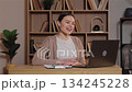 Caucasian woman smiling and waving hand during online meeting on laptop. Female student attending virtual class sitting at wooden desk in cozy home study area with shelves full of books behind. 134245228