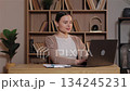 Caucasian woman sitting at wooden desk opening laptop and beginning work. Female preparing to type starting online task in calm home office with books arranged on wooden shelves behind her. 134245231