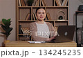 Caucasian woman looking at camera smiling while sitting at desk and typing on laptop. Female working from home feeling satisfied with progress in calm workspace with wooden shelves and plants behind. 134245235