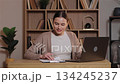 Caucasian woman smiling while working at wooden desk using laptop and reviewing notes. Female entering data from documents sitting in cozy home office with shelves full of books in background. 134245237