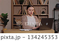 Caucasian woman analyzing charts on printed report holding pen and typing on laptop. Female checking financial data entering information at wooden desk in home office with bookshelf background. 134245353