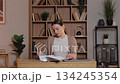 Caucasian woman sitting at wooden desk reading papers and marking notes. Female concentrating on work reviewing documents at home workspace with laptop and organized shelves filled with books behind. 134245354