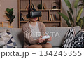 Caucasian woman wearing VR headset leaning forward holding controller. Female immersed in action video game dodging virtual obstacle at home sitting on couch in room with wooden shelves. 134245355