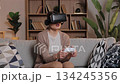 Caucasian woman wearing VR headset holding game controller sitting on couch. Female enjoying virtual reality videogame experience at home in cozy living room with wooden shelves behind. 134245356