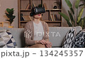 Caucasian woman wearing virtual reality headset sitting on sofa at home interacting with digital space. Female raising hands gesturing to magnify virtual object in relaxed room with shelves. 134245357