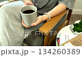 A woman sits in a gray chair, drinking coffee beside a white book-filled table, creating a serene blend of comfort, warmth, and minimalist home style. 134260389