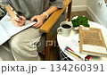 A woman takes notes in a notebook on a gray armchair next to a white table with books and coffee, forming a peaceful workspace moment. 134260391