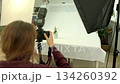 A woman in red photographs a souvenir in a bright minimalist studio, using softboxes to create a clean, professional product-shoot atmosphere. 134260392