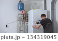 Technician installing indoor heat pump system with pipes and expansion tank. Man adjusting control panel on heat pump. Concept of home heating installation, energy efficiency and HVAC maintenance 134319045