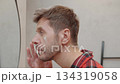 Man in red pajamas applying face cream while looking at mirror in modern bathroom. Concept of male skincare, grooming, self care and daily hygiene 134319058