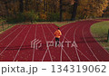 Man running on red track in autumn park. Man in orange sportswear jogging at sport stadium with fallen autumn leaves. Concept of fitness, wellness and outdoor training 134319062