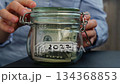 Female hands turn Glass jar full of American currency dollars cash banknote with text 2027 year. Preparation saving money. Moderate consumption and economy 134368853