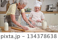 Happy family in kitchen. Grandmother and granddaughter child cook in kitchen together. Grandma teaching kid girl knead dough bake cookies. Household teamwork helping family generations concept 134378638